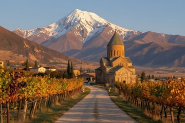 Obraz premium Ancient church stands in vineyard landscape with snowy mountain peak. Golden autumn foliage lines road leading to village. Serene rural scene at sunset.