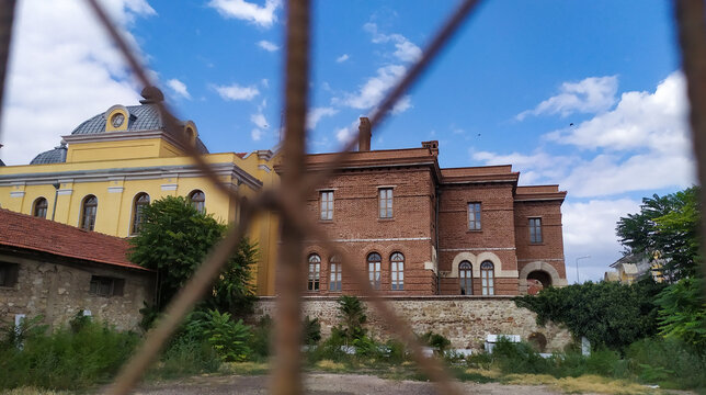 Historic Edirne Synagogue complex seen through iron fence, yellow facade and red brick annex, Turkey. Abandoned Jewish heritage site.