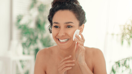A woman is smiling while using a cotton pad to apply skincare product on her face. The setting is a bright bathroom with natural light. This activity is part of her daily morning routine. © Prostock-studio