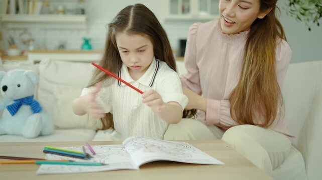 Affectionate Asian mother gently touches her little daughter's hair as the sweet girl focuses on coloring a book, enjoying cozy family bonding time in a bright, modern living room at home.