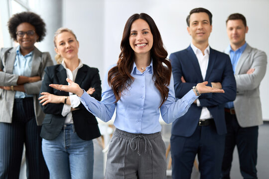 A smiling young businesswoman stands in front of her colleagues with open arms, welcoming gesture, smiling confidently at the camera. Four diverse colleagues stand behind her in bright modern office