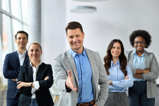 Smiling businessman offering a handshake in a modern office. His colleagues are in the background