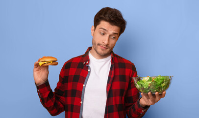A young man stands against a blue wall. He holds a burger in one hand and a bowl of salad in the other hand. His expression shows he is thinking about which food to choose. © Prostock-studio