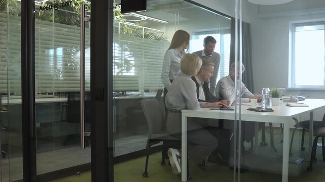 Group of business people standing together and discussing their work and projects, having a team meeting in an office, view from glass wall