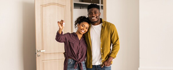 A young couple stands in their new home, smiling and holding keys. They are in a bright room with light walls and a wooden door. The space looks empty, indicating a fresh start. © Prostock-studio