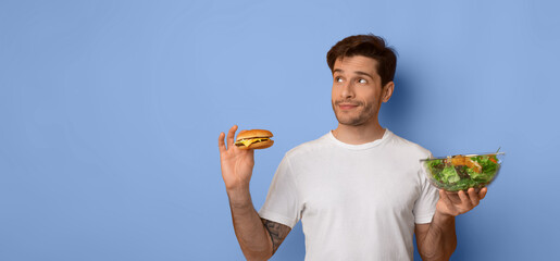 A man stands in front of a blue wall holding a burger in one hand and a bowl of salad in the other. He looks thoughtful about what to eat next. © Prostock-studio