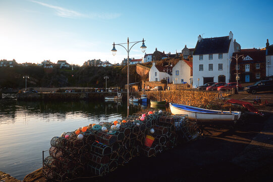 Quiet waterfront harbor with fishing boats, lobster pots, stone walls, and village houses in warm evening light under a clear sky. Crail, Scotland