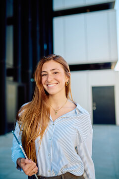 Young woman with red hair and freckles smiling, standing outdoors holding a digital tablet with a modern office building