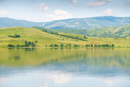 Green mountains and the blue sky with clouds are reflected in the lake. Beautiful summer landscape at sunny day.