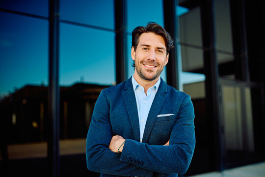 Young professional man with a confident smile and crossed arms standing in front of a modern glass office building