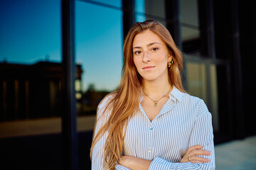 Fototapeta premium Young businesswoman with freckles and long red hair standing outdoors with crossed arms, looking at camera