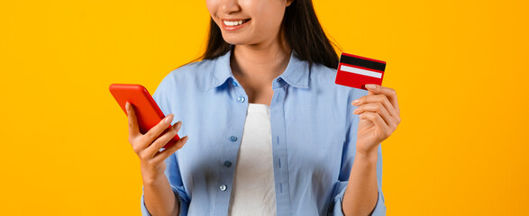 A person stands against a yellow background holding a smartphone in one hand and a credit card in the other. She is smiling and appears engaged in an activity with the devices. © Prostock-studio