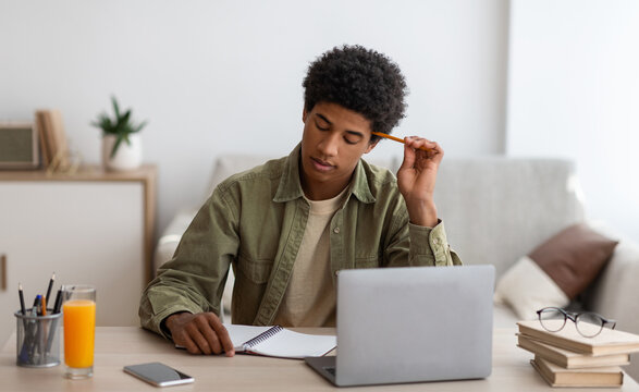 Thoughtful black teen guy passing hard exam or online test on laptop at home, panorama. Teenage student learning remotely, doing difficult home assignment, working on educational project