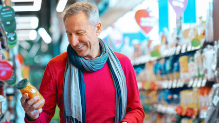 A man stands in a grocery store holding a can of food and smiling. He is dressed in a red sweater and grey scarf. Shelves full of products are visible around him. © Prostock-studio