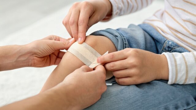 A person is applying a fabric adhesive bandage to a child's knee, indicating care and first aid for a minor injury or wound.