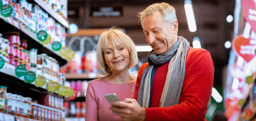 A couple stands in a grocery store looking at a phone together. They smile as they browse items on shelves filled with products. They appear engaged with their shopping experience. © Prostock-studio