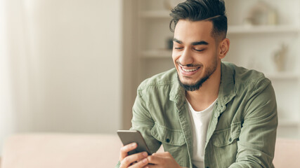 A young man sits on a couch at home, looking at his smartphone with a smile. He is dressed casually...