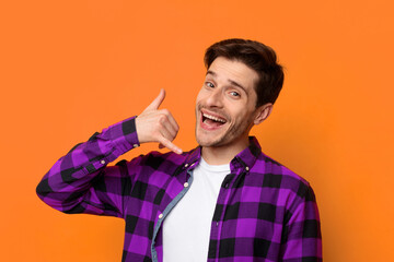 A man stands in front of an orange background. He is smiling and making a calling gesture with his hand. The scene shows a cheerful and lively moment. © Prostock-studio