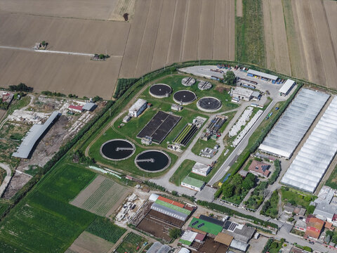 Aerial view of a water treatment plant with circular sedimentation tanks and rectangular basins surrounded by green fields and greenhouses in Paestum, Campania, Italy.