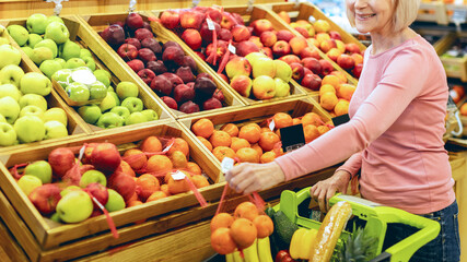 A woman is selecting fruits and vegetables at a market. She stands next to wooden bins filled with colorful apples and oranges. She holds a green basket with items. © Prostock-studio