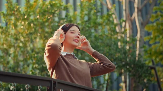 Young Asian woman in a brown sweater putting on wireless headphones to enjoy music at a green park during golden hour. Serene female professional relaxing outdoors with podcast or audio.
