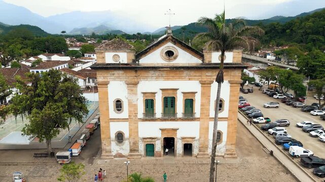 Aerial view overflying First Church with colonial rooftops, mountains and main square in historic Paraty, Brazil. Push in, pan and overfly camera movement