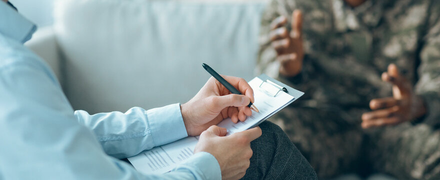 A counselor sits with a clipboard in hand, writing notes while engaging in conversation with a service member. The focus is on sharing experiences and emotions in a confidential atmosphere.