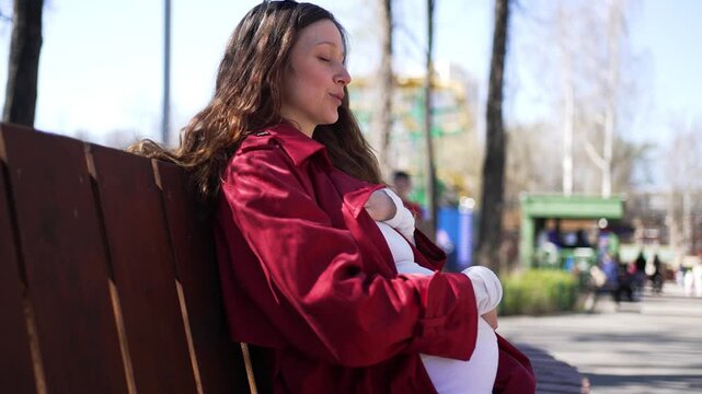 Content pregnant woman relaxing on a park bench during a beautiful sunny day, she gently caresses her belly while enjoying the pleasant weather and the outdoor atmosphere