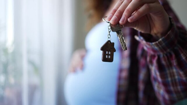 Expectant mother holding keys with a house keychain, her other hand gently resting on her pregnant belly, embodying the concept of family growth and acquiring a new home