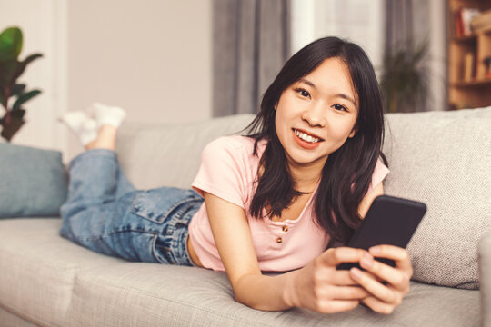 A young woman relaxes on a couch in a living room. She lies on her stomach with her feet in the air. She holds a smartphone and smiles while looking at the screen.