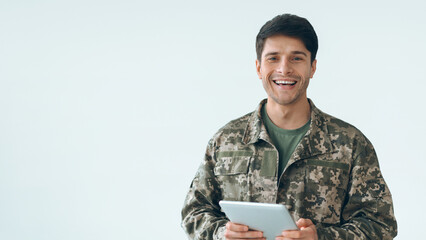 A soldier stands indoors at a military base, smiling while holding a tablet. The soldier is wearing a camouflage uniform. The background is plain and light-colored. © Prostock-studio