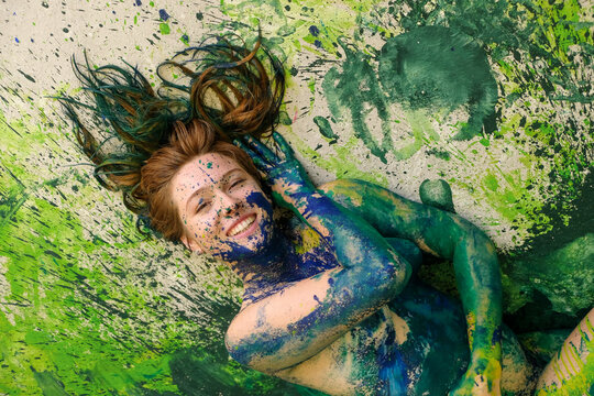 Smiling woman covered in vivid blue and green paint lying on splattered surface creating an energetic artistic portrait filled with movement expression and creative freedom