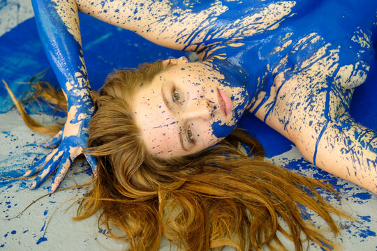 Red haired woman covered in vivid blue paint lying in an expressive artistic composition with dynamic splashes, creating a bold fusion of body art and emotion