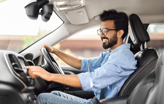 Road playlist. Side view of happy young indian man driving car and listening to music radio on auto audio system, checking automobile options during test drive, pushing button on dashboard