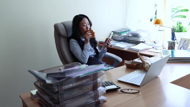 Asian woman worker in office having fast food lunch at desk. Woman eating hamburger while working in office in front of the computer.