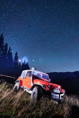 Fototapeta premium Red off road jeep parked on grassy mountain slope at night, starry sky with Milky Way above forest silhouettes, dramatic low angle wilderness scene: Bukovel, Ukraine - August 15, 2023