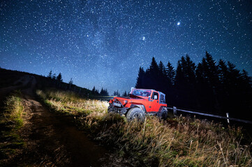 Fototapeta premium Off road jeep parked by dirt track under starry sky, Milky Way above mountain forest, concept of night journey, exploration and remote adventure: Bukovel, Ukraine - August 15, 2023