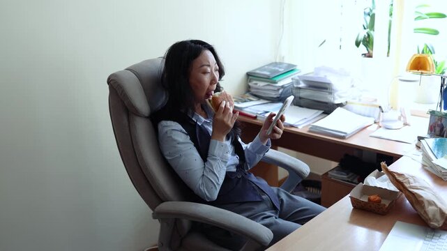 Asian woman worker in office having fast food lunch at desk. Woman eating hamburger while working in office in front of the computer.