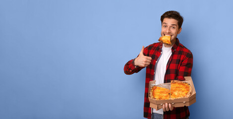 A man stands in front of a blue background. He holds a pizza box with one slice in his mouth. He smiles and gives a thumbs up with his free hand. © Prostock-studio