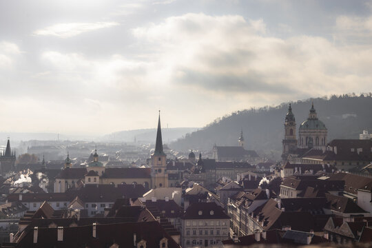View of historic red rooftops and church spires under a hazy morning sky with Petrin Hill in the background in Prague, Prague, Czechia.