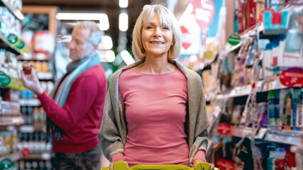A woman stands in a grocery store holding a shopping cart. She smiles while looking at the camera. In the background, a man browses products on the shelf. The store is well stocked. © Prostock-studio