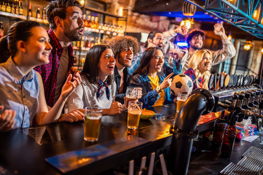 Diverse friends cheering for soccer match with beer at bar counter.