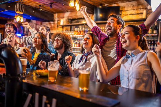 Excited friends shouting and celebrating soccer goal with beer in a pub.