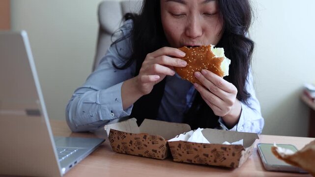 Asian woman worker in office having fast food lunch at desk. Woman eating hamburger while working in office in front of the computer.