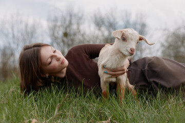 Fototapeta premium Lifestyle scene with a woman lying on lush grass, a goat kid nearby, soft light, candid expression, natural colors, peaceful outdoors, gentle affection and connection in a rural meadow