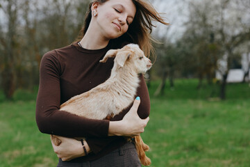Fototapeta premium Joyful woman outdoors holding a goat kid in her arms, park setting, casual sweater, candid lifestyle moment, natural light, warm mood, gentle bond with animal, authentic and serene countryside scene