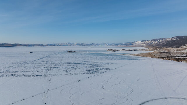 Wide winter view of the frozen Small Sea strait on Lake Baikal with snow-covered ice, distant islands and mountain shoreline under clear blue sky, Russia