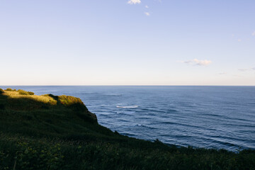 Naklejka premium Wide seascape view from a grassy coastal cliff with gentle ocean waves and a clear sky, ideal for travel and nature backgrounds with copy space