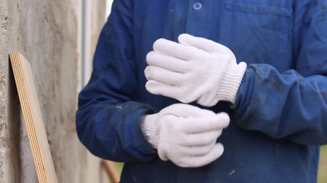 A builder puts on white work gloves before starting work. A master in blue overalls puts on protective knitted gloves on a construction site background. Close-up of a person at work