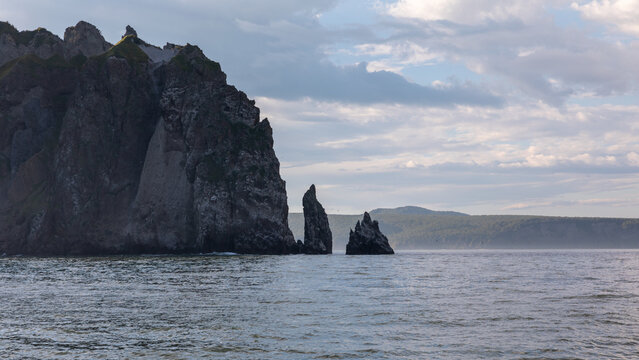 Rugged coastal cliffs and sharp sea stacks rising from calm water in Avacha Bay, Kamchatka Krai, Russia, under dramatic cloudy sky at dusk